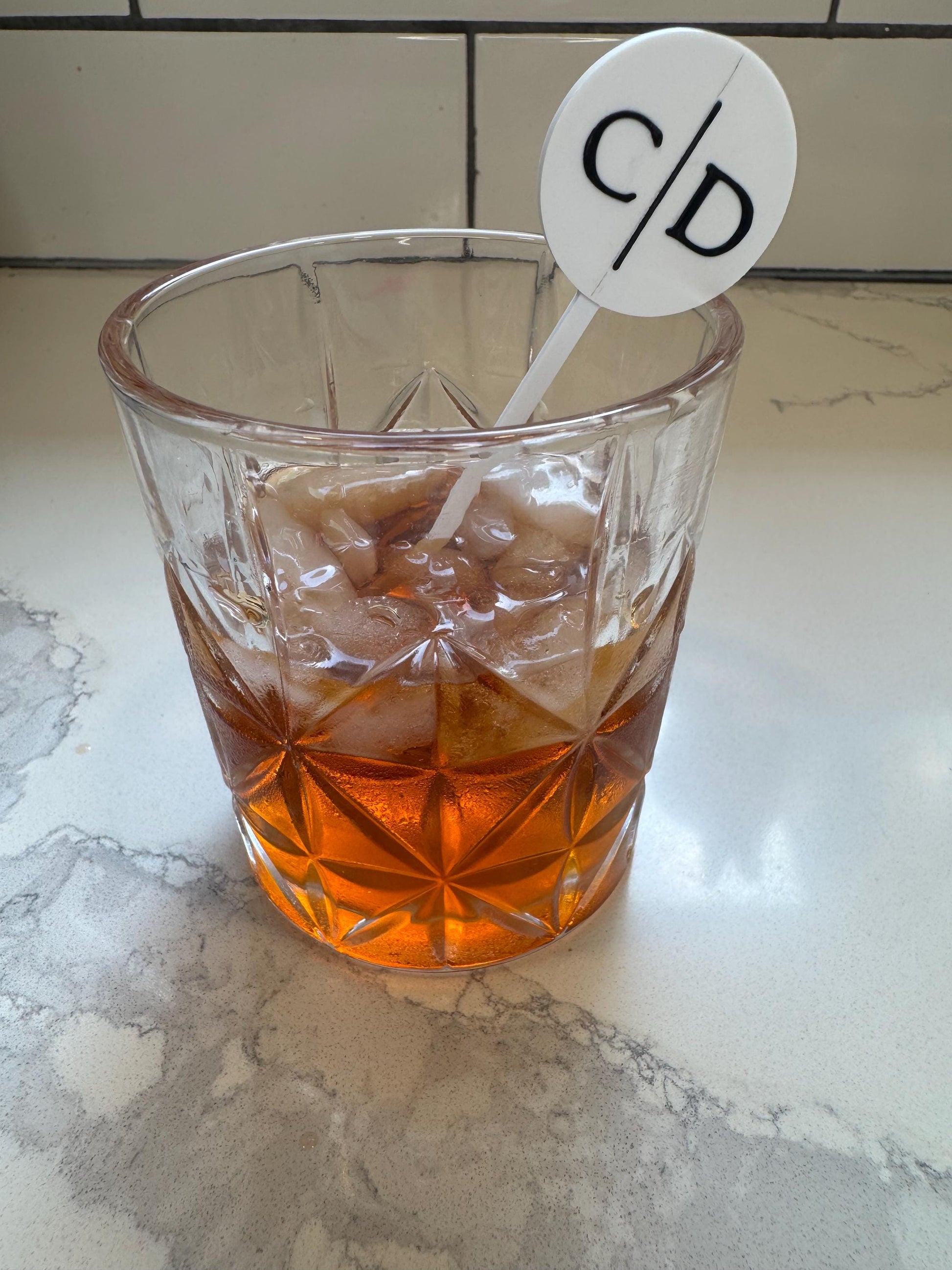 May include: A clear, faceted glass tumbler filled with amber liquid and ice cubes, featuring a white circular drink stirrer with a thin vertical line separating the black letters C and D, positioned upright inside the glass.
