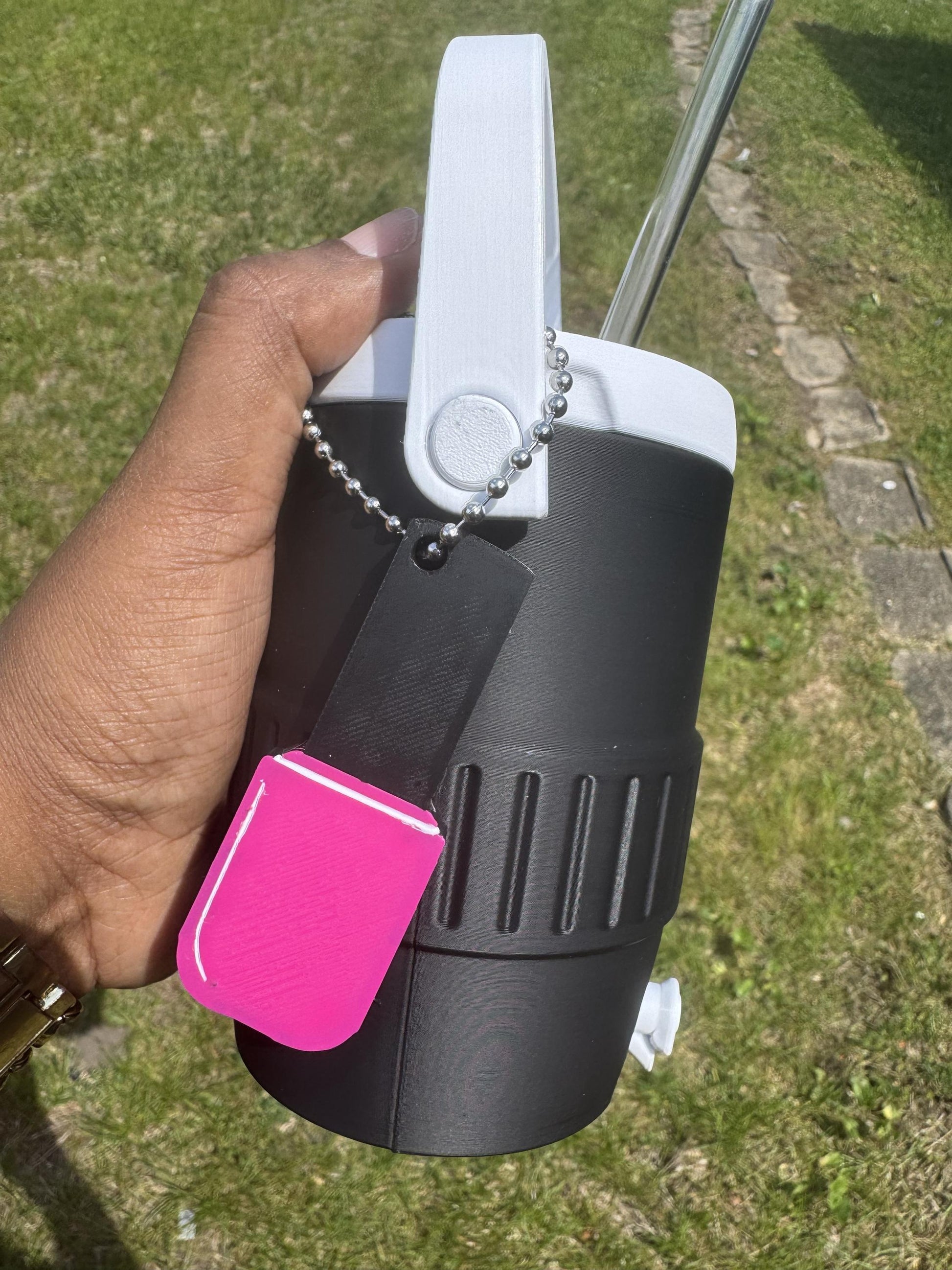 May include: A black insulated beverage cooler with a white handle and a silver metal straw. A black and bright pink rectangular tag hangs from the handle by a silver ball chain. The cooler features a textured grip section and a white spout.