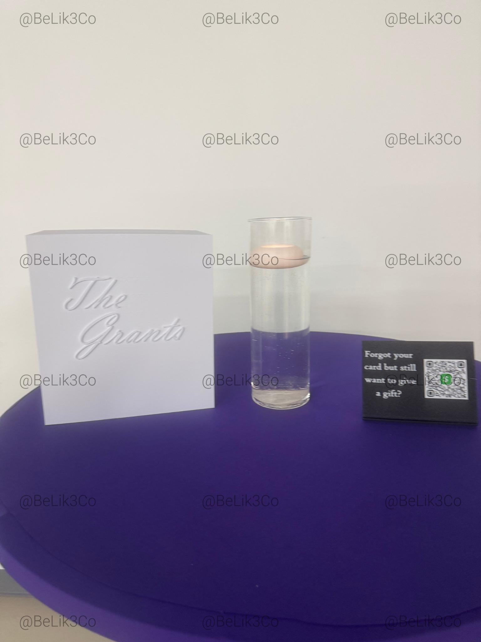 May include: A white square box with the embossed text The Grants, a clear cylindrical glass vase containing water and a floating light pink candle, and a small black rectangular sign with white text and a green QR code icon.