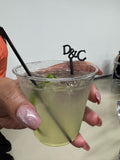 May include: A clear plastic cup filled with a pale yellow liquid, ice cubes, and a lime wedge. A black straw and a black decorative stirrer featuring the letters D&C are inserted into the drink. The cup has a wide rim and a tapered base design.