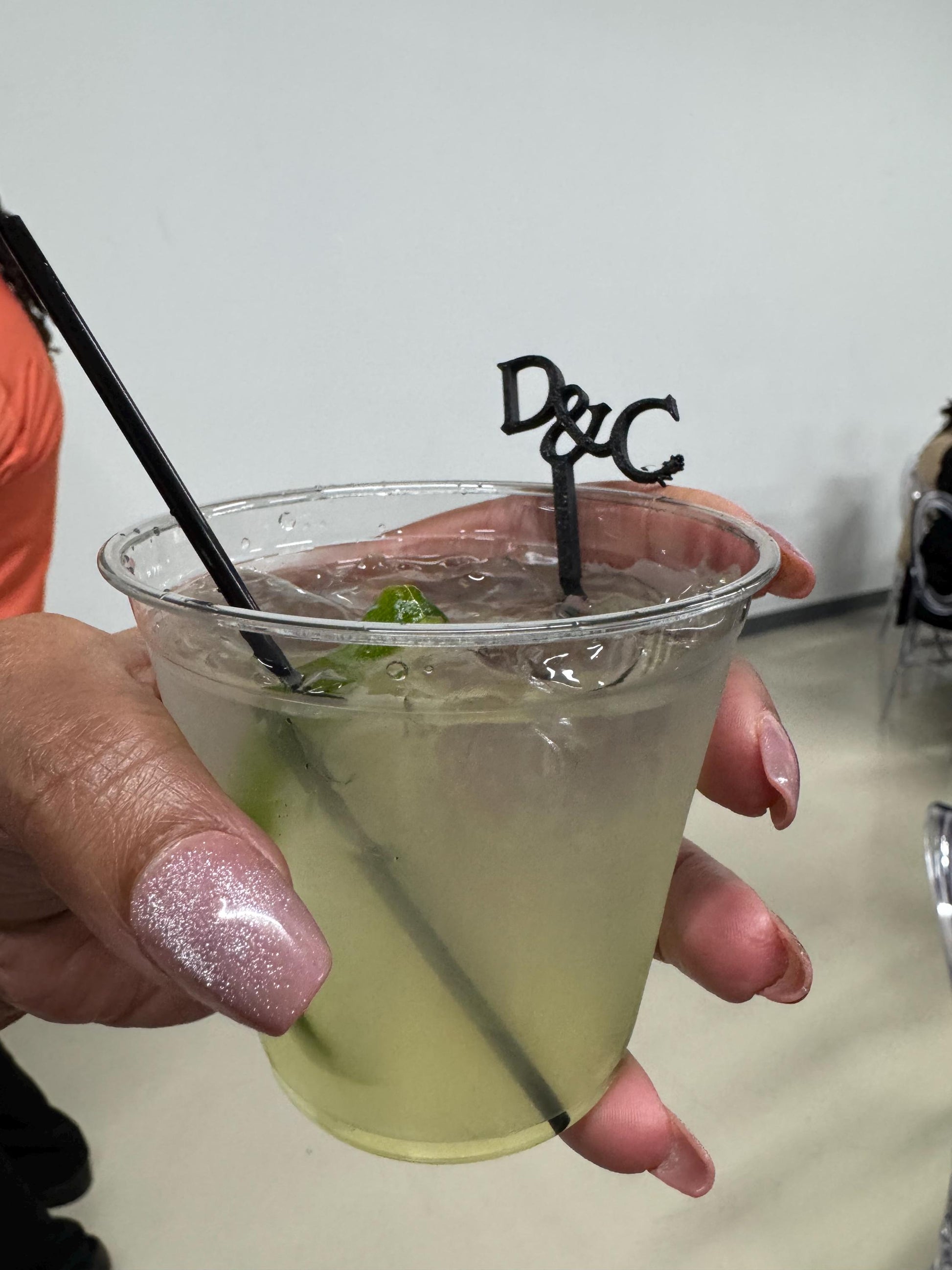 May include: A clear plastic cup filled with a pale yellow liquid, ice cubes, and a lime wedge. A black straw and a black decorative stirrer featuring the letters D&amp;C are inserted into the drink. The cup has a wide rim and a tapered base design.