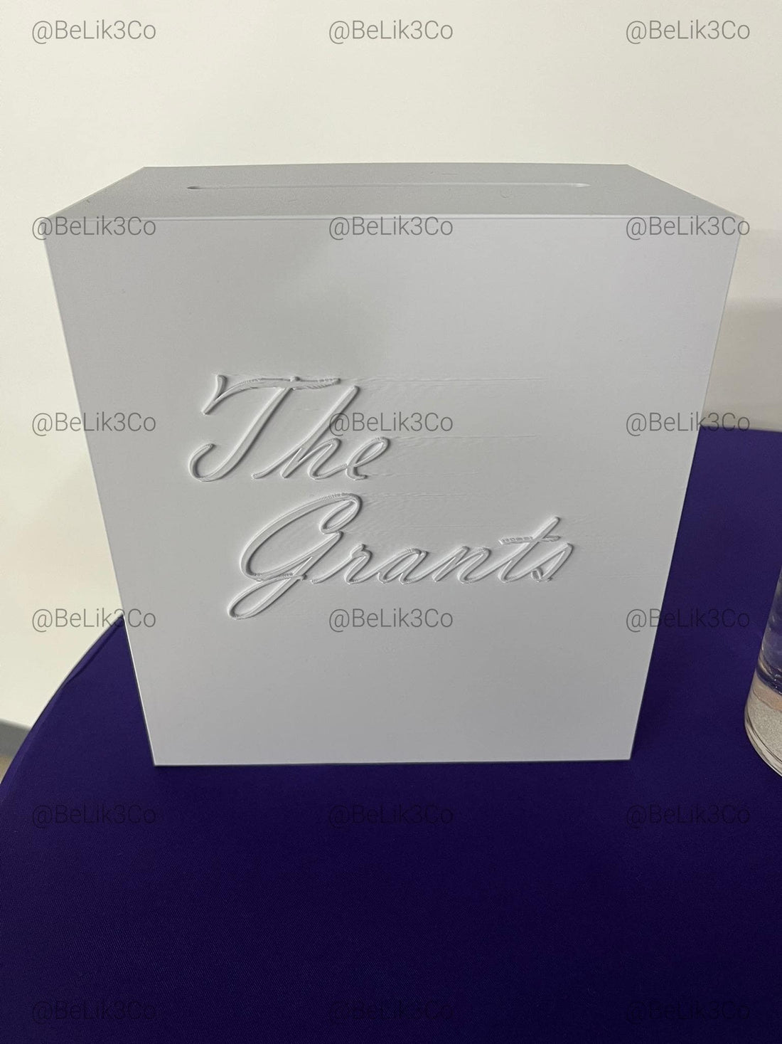 May include: A white rectangular sign with the black text The Grants, a clear cylindrical glass vase containing water and a white floating candle, and a small black rectangular sign with white text and a QR code for gift giving.